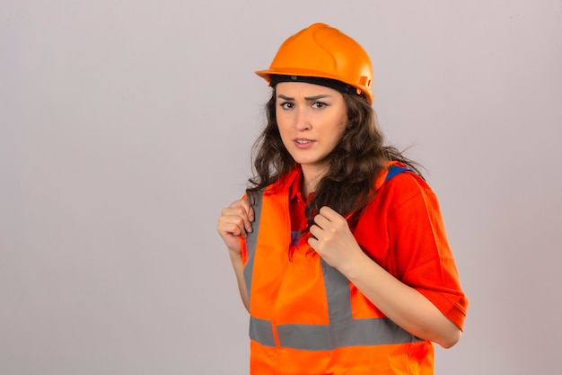 Mulher jovem construtor em uniforme de construção e capacete de segurança, franzindo a testa com expressão cética de dúvida sobre a parede branca isolada