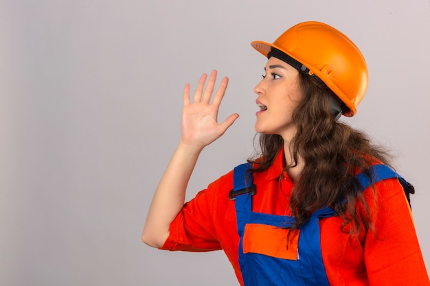 Mulher jovem Construtor em uniforme de construção e capacete de segurança, gritando para o lado com a mão na boca sobre parede branca isolada