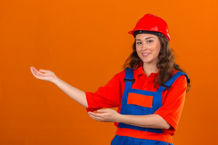 Foto grátis mulher jovem construtor em uniforme de construção e capacete de segurança sorrindo alegre apresentando e apontando com as palmas das mãos, olhando para a câmera sobre parede laranja isolada