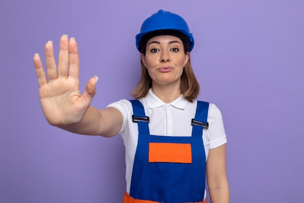 Mulher jovem construtora com uniforme de construção e capacete de segurança com cara séria, fazendo gesto de parada com a mão em pé sobre a parede roxa