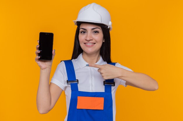 Mulher jovem construtora com uniforme de construção e capacete de segurança mostrando o smartphone apontando com o dedo indicador para ele e sorrindo alegremente em pé sobre a parede laranja