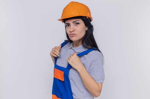 Mulher jovem construtora com uniforme de construção e capacete de segurança, olhando para a frente com expressão séria e confiante em pé sobre a parede branca