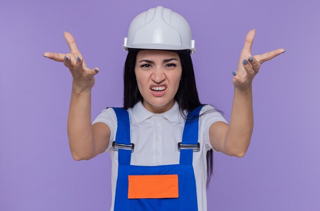 Mulher jovem construtora com uniforme de construção e capacete de segurança olhando para a frente com os braços levantados, como se estivesse discutindo em pé sobre a parede roxa