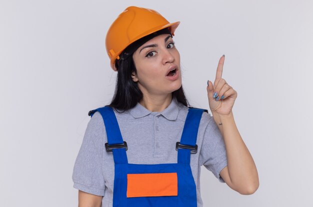 Mulher jovem construtora com uniforme de construção e capacete de segurança olhando para a frente surpresa, mostrando o dedo indicador tendo uma nova boa ideia em pé sobre uma parede branca