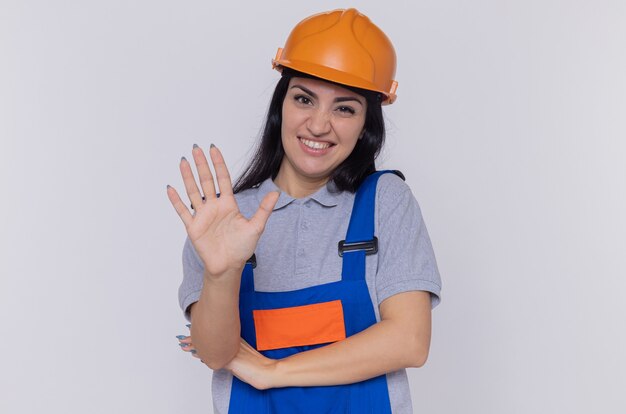 Mulher jovem construtora com uniforme de construção e capacete de segurança, olhando para frente com um sorriso cético no rosto, mostrando a palma da mão aberta sobre a parede branca