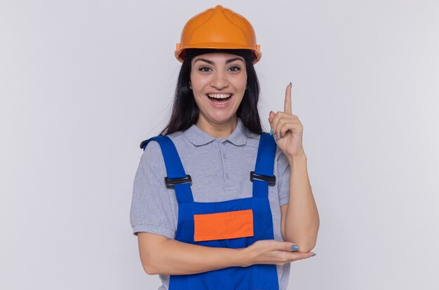 Mulher jovem construtora com uniforme de construção e capacete de segurança, olhando para frente com um sorriso no rosto inteligente, mostrando o dedo indicador em pé sobre uma parede branca