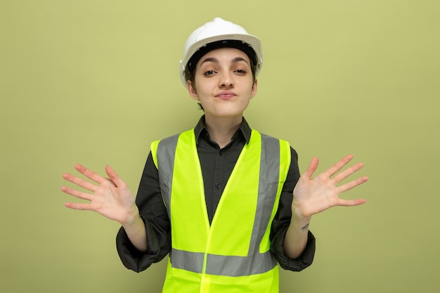 Foto grátis mulher jovem construtora em colete de construção e capacete de segurança confusa abrindo os braços para os lados em pé no verde