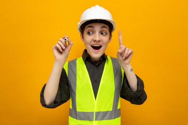 Mulher jovem construtora em colete de construção e capacete de segurança parecendo surpresa sorrindo, mostrando o dedo indicador tendo uma ótima ideia escrevendo com uma caneta no ar em pé sobre a parede laranja