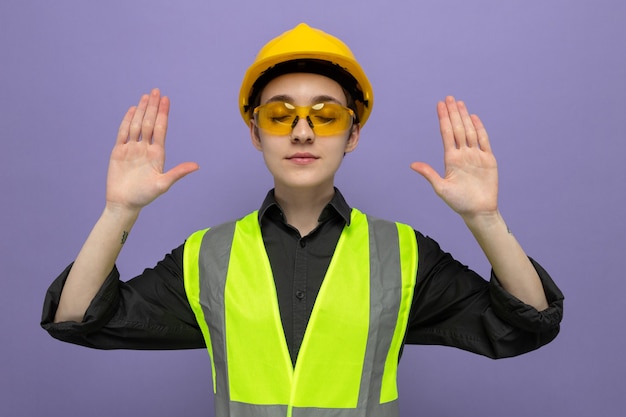 Mulher jovem construtora em colete de construção e capacete de segurança usando óculos de segurança amarelos, feliz e positiva levantando os braços