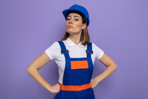 Mulher jovem construtora em uniforme de construção e capacete de segurança com expressão séria e confiante com os braços na altura do quadril em pé no roxo