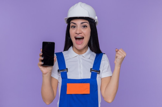 Mulher jovem construtora em uniforme de construção e capacete de segurança mostrando smartphone olhando para frente feliz e animada com o punho cerrado em pé sobre a parede roxa