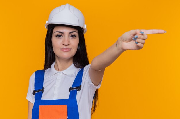 Mulher jovem construtora em uniforme de construção e capacete de segurança olhando para a frente sorrindo confiante apontando com o dedo indicador para o lado em pé sobre a parede laranja