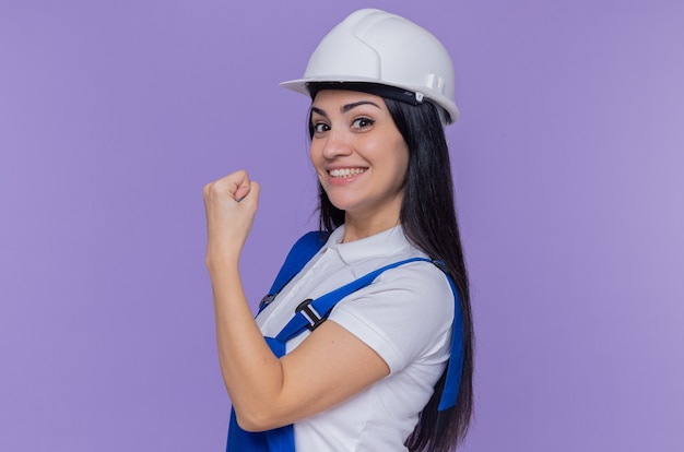 Mulher jovem construtora em uniforme de construção e capacete de segurança, olhando para a frente, sorrindo confiante, mostrando o punho em pé sobre a parede roxa