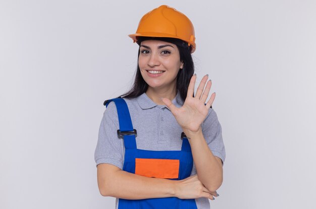 Mulher jovem construtora em uniforme de construção e capacete de segurança olhando para frente feliz e positiva, mostrando a palma da mão aberta sobre a parede branca