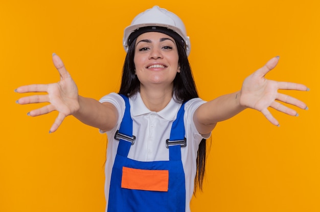 Foto grátis mulher jovem construtora em uniforme de construção e capacete de segurança olhando para frente sorrindo alegremente fazendo gesto de boas-vindas e abrindo as mãos em pé sobre a parede laranja