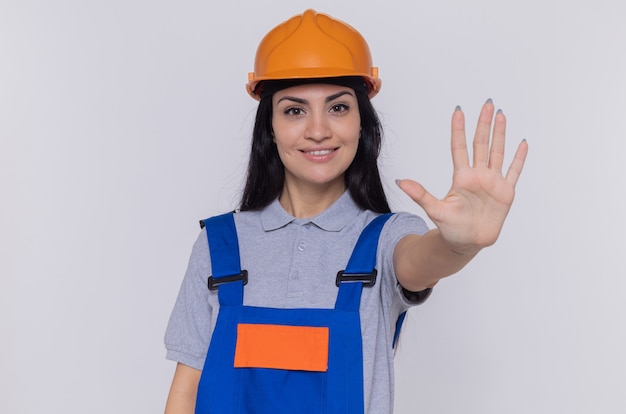 Mulher jovem construtora em uniforme de construção e capacete de segurança olhando para frente sorrindo confiante, fazendo gesto de parada com a mão aberta em pé sobre a parede branca