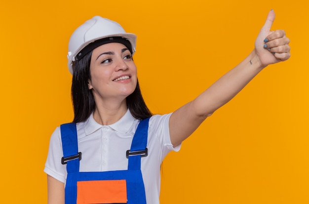 Mulher jovem construtora em uniforme de construção e capacete de segurança olhando para o lado e sorrindo confiante mostrando o polegar em pé sobre a parede laranja