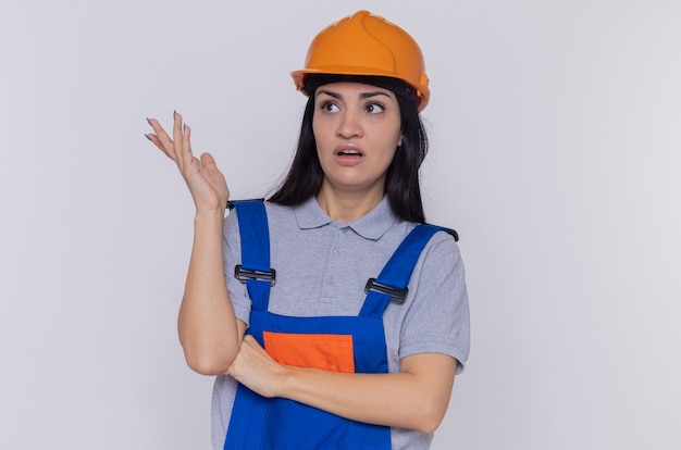 Mulher jovem construtora em uniforme de construção e capacete de segurança, parecendo confusa e descontente com o braço estendido em pé sobre uma parede branca