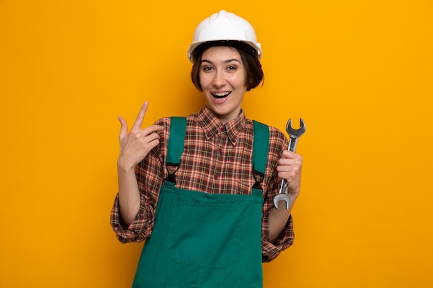 Mulher jovem construtora em uniforme de construção e capacete de segurança segurando a chave, feliz e surpresa, mostrando o dedo indicador em pé na laranja