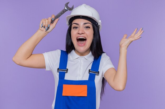 Mulher jovem construtora em uniforme de construção e capacete de segurança segurando a chave inglesa, levantando o braço, gritando feliz e animada em pé sobre a parede roxa