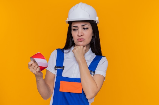 Mulher jovem construtora em uniforme de construção e capacete de segurança segurando fita adesiva olhando para ela com a mão no queixo pensando em pé sobre a parede laranja