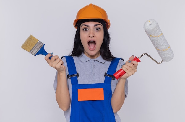 Foto grátis mulher jovem construtora em uniforme de construção e capacete de segurança segurando rolo de pintura e escova olhando para frente surpresa em pé sobre uma parede branca