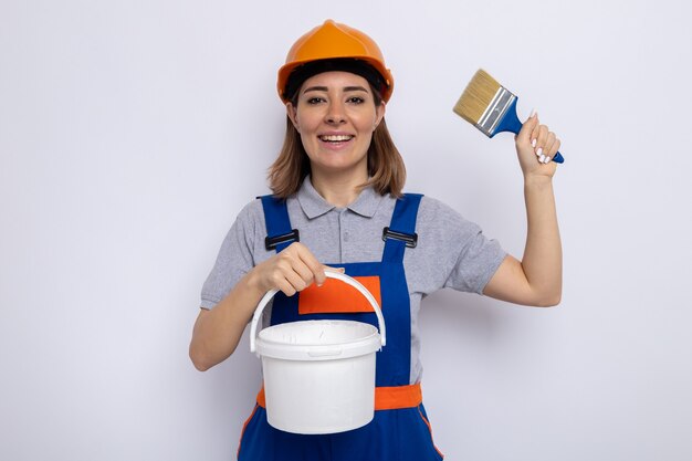 Mulher jovem construtora em uniforme de construção e capacete de segurança segurando um balde de tinta e uma escova pait feliz e positiva sorrindo alegremente em pé sobre uma parede branca