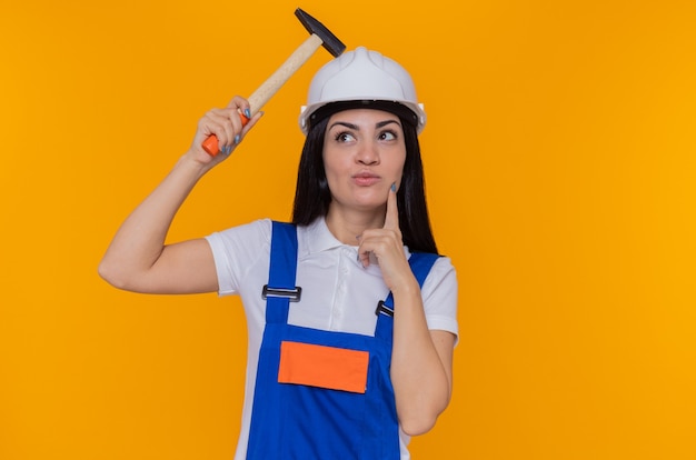 Mulher jovem construtora em uniforme de construção e capacete de segurança segurando um martelo, olhando para cima com uma expressão pensativa no rosto pensando em pé sobre a parede laranja
