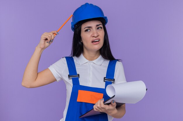 Mulher jovem construtora em uniforme de construção e capacete de segurança segurando uma prancheta e um lápis, parecendo confusa e coçando a cabeça em pé sobre a parede roxa