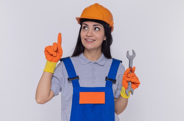 Mulher jovem construtora em uniforme de construção e capacete de segurança usando luvas de borracha segurando uma chave inglesa mostrando o dedo indicador olhando para cima sorrindo em pé sobre uma parede branca