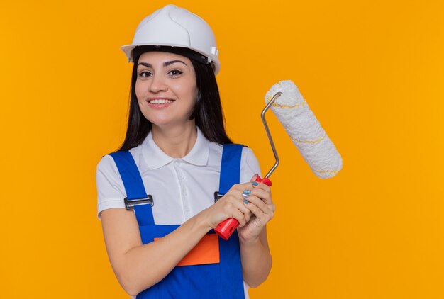 Mulher jovem construtora feliz com uniforme de construção e capacete de segurança segurando o rolo de pintura, olhando para a frente, sorrindo confiante em pé sobre a parede laranja