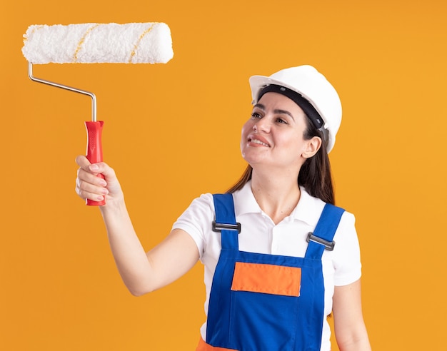 Mulher jovem construtora sorridente de uniforme levantando e olhando para a escova de rolo isolada na parede laranja