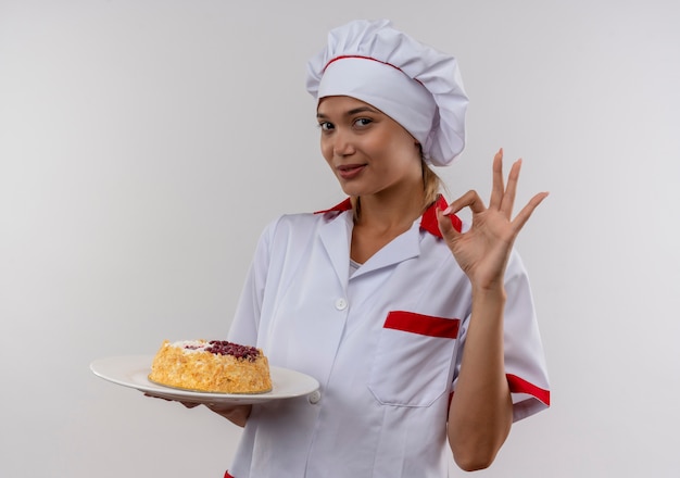 Foto grátis mulher jovem cozinheira satisfeita vestindo uniforme de chef segurando bolo no prato, mostrando gesto ok na parede branca isolada com espaço de cópia