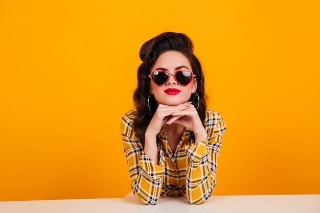 Mulher jovem curiosa sentada sobre fundo amarelo. Foto de estúdio de linda garota Pin-up usando óculos em forma de coração.