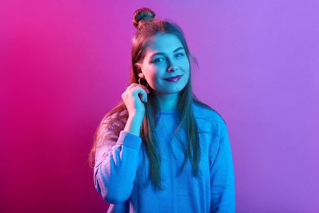 Mulher jovem de aparência agradável usa um suéter casual, tem coque no cabelo, parece com expressões faciais calmas, posa contra um espaço neon rosa