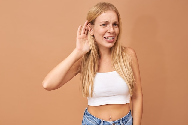 Mulher jovem de blusa branca e calça jeans com a mão na orelha, ouvindo com atenção