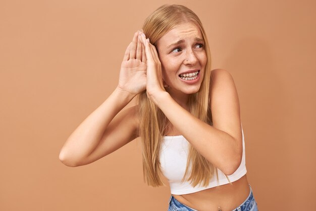 Mulher jovem de blusa branca e calça jeans com a mão na orelha, ouvindo com atenção