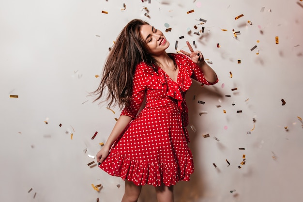 Foto grátis mulher jovem de bom humor está usando um vestido de bolinhas vermelhas. menina com sorriso posa na parede de confete e mostra o símbolo da paz.