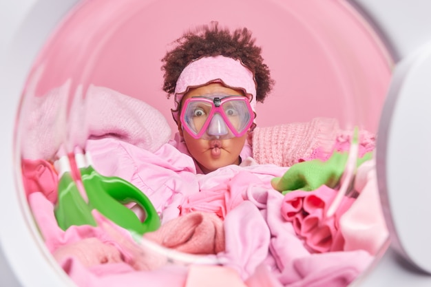 Mulher jovem de cabelo encaracolado engraçada faz careta lábios de peixe usa máscara de mergulho em poses de dentro da máquina de lavar se prepara para o processo de lavagem cercada por uma pilha de roupas sujas para lavar