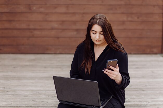 Mulher jovem de cabelos castanhos com laptop e computador telefone inteligente, sentada no chão de madeira