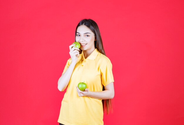 Mulher jovem de camisa amarela segurando uma maçã verde e dando uma mordida