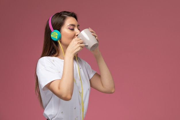 Mulher jovem de camisa branca bebendo café e ouvindo música na parede rosa modelo mulher de frente