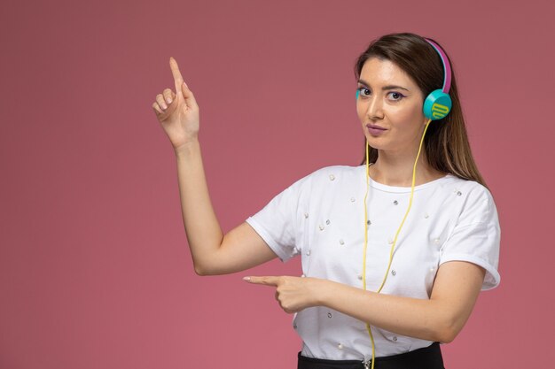 Mulher jovem de camisa branca posando de frente para ouvir música com os fones de ouvido na parede rosa.
