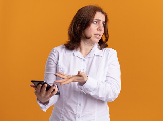 Mulher jovem de camisa branca segurando um smartphone, apontando com o braço para ele, estando confusa e descontente em pé sobre a parede laranja