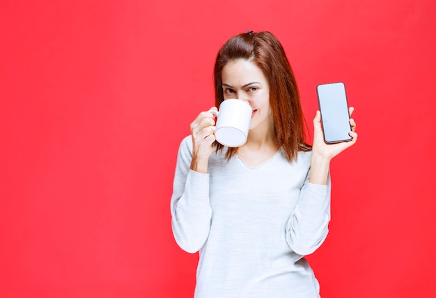 Mulher jovem de camisa branca segurando uma caneca de café branca e um smartphone preto