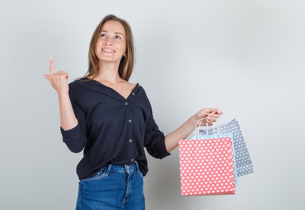 Mulher jovem de camisa preta, shorts jeans apontando para cima com sacos de papel e parecendo alegre