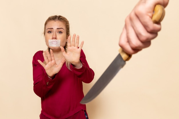 Mulher jovem de camisa vermelha com a boca amarrada de frente com medo de faca no espaço creme feminino pano foto violência doméstica