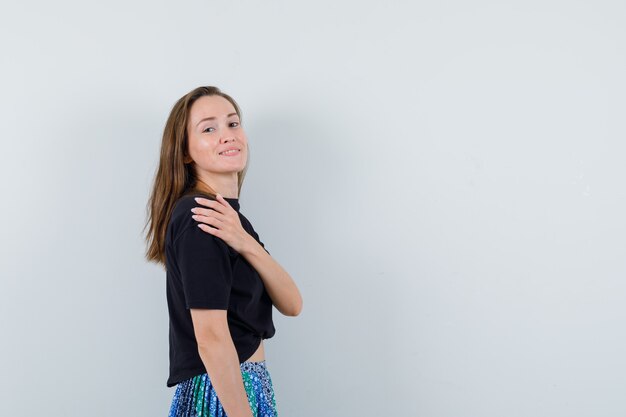 Mulher jovem de camiseta preta e saia azul em pé, olhando por cima do ombro e parecendo feliz