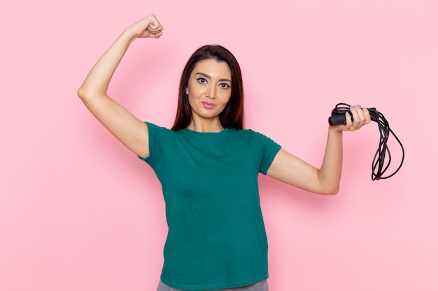 Mulher jovem de camiseta verde segurando a corda de pular e flexionando-se na parede rosa claro cintura esporte exercícios exercícios beleza atleta feminina de frente