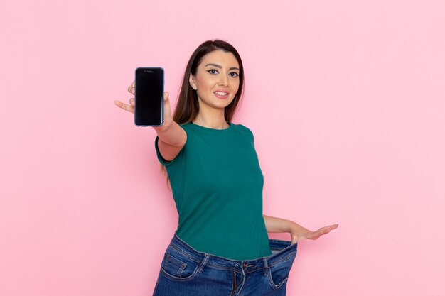 Mulher jovem de camiseta verde segurando o smartphone na parede rosa claro cintura exercício treino de beleza esporte feminino